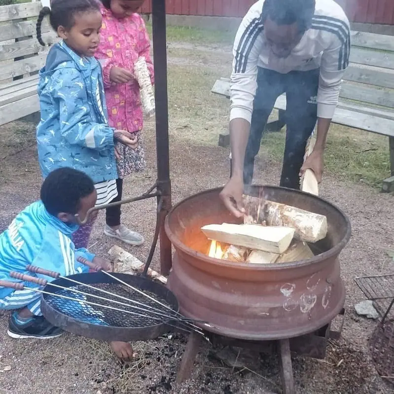 Lapset ja aikuinen kokoontuvat ulkona tulisijan ympärille.